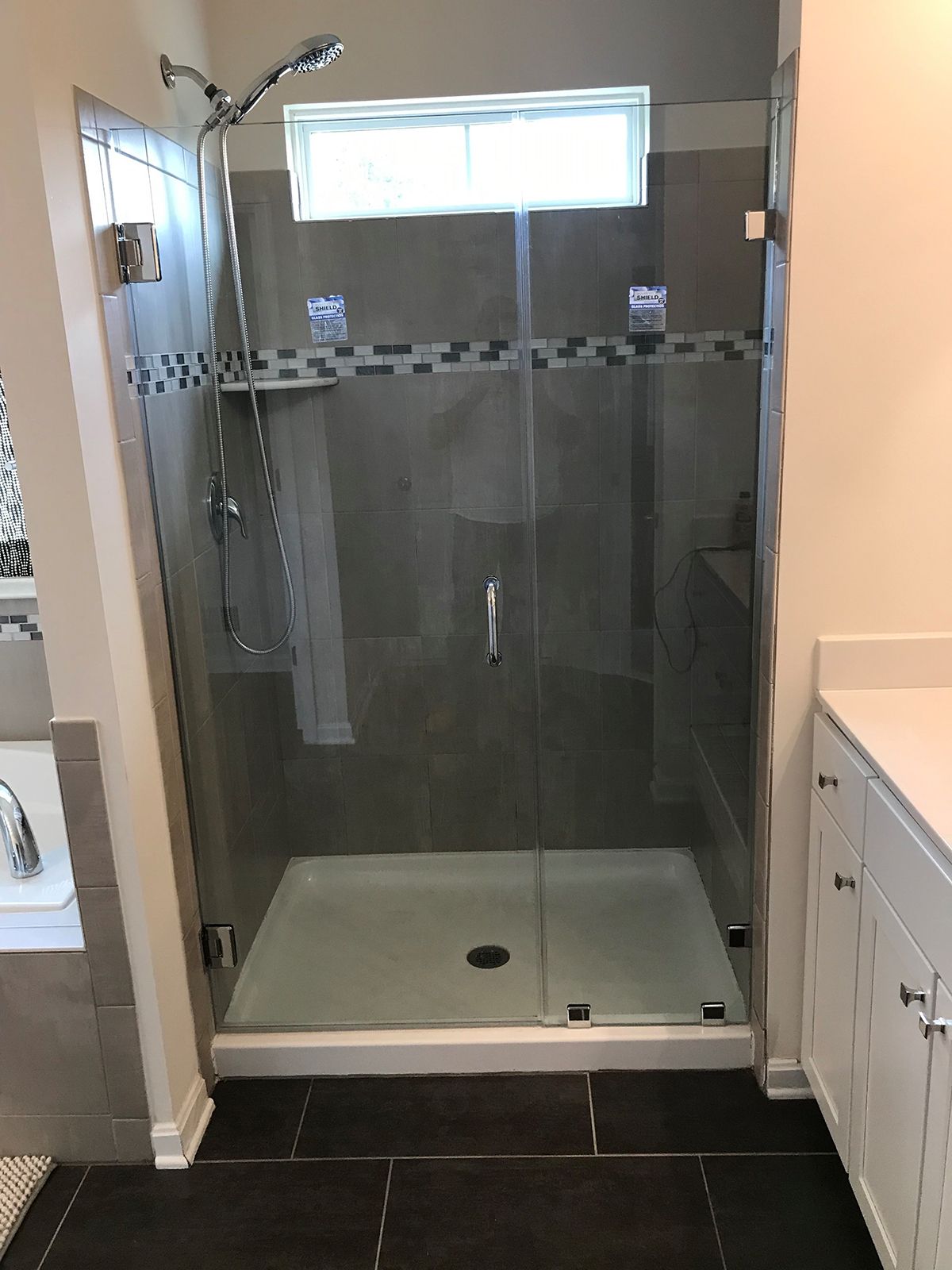 A walk-in shower with a glass door, light grey tile walls, a horizontal decorative accent border, and a white shower pan.