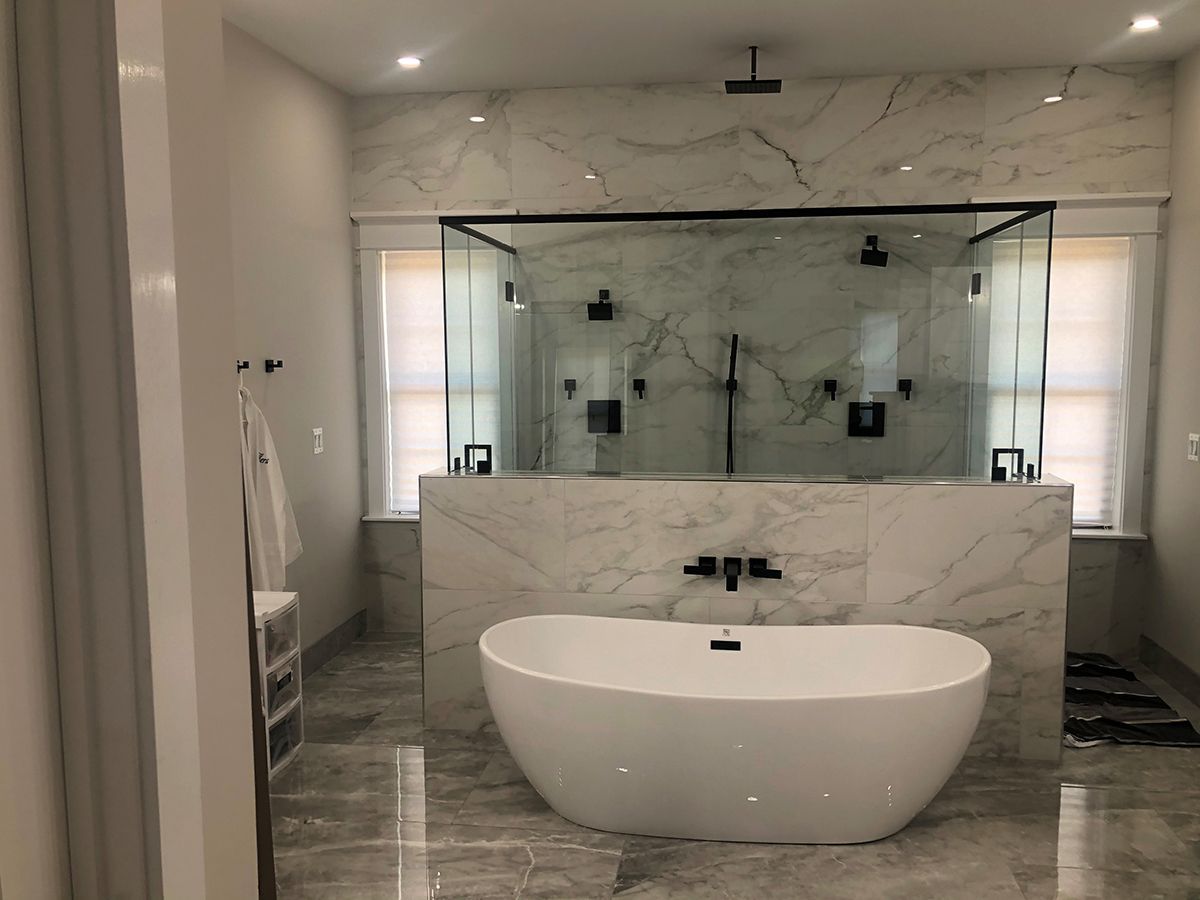 A freestanding white bathtub sits in front of a glass-enclosed shower with marble tile walls in a modern bathroom.