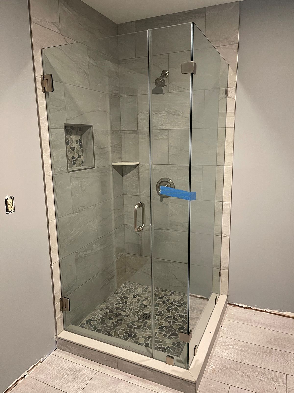 A modern glass shower enclosure with gray stone-look tiles and a pebble floor inside a room with gray walls.