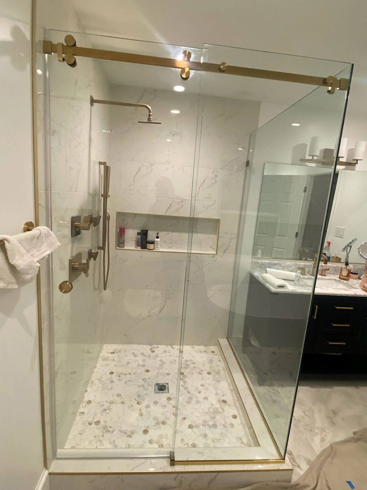 A modern walk-in shower with gold fixtures, a glass sliding door, marble tile walls, and a pebble-stone floor.