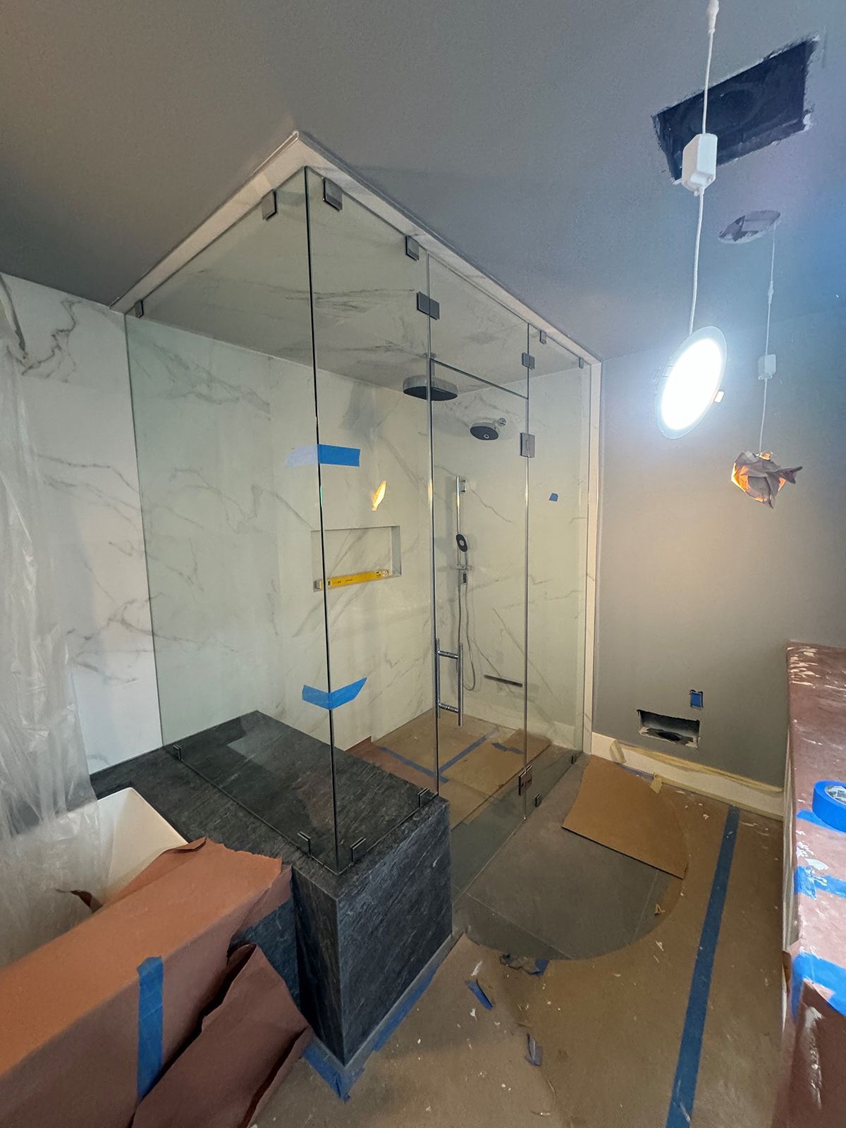 A partially finished bathroom featuring a glass-enclosed shower with marble-look tile, dark flooring, and hanging lights.