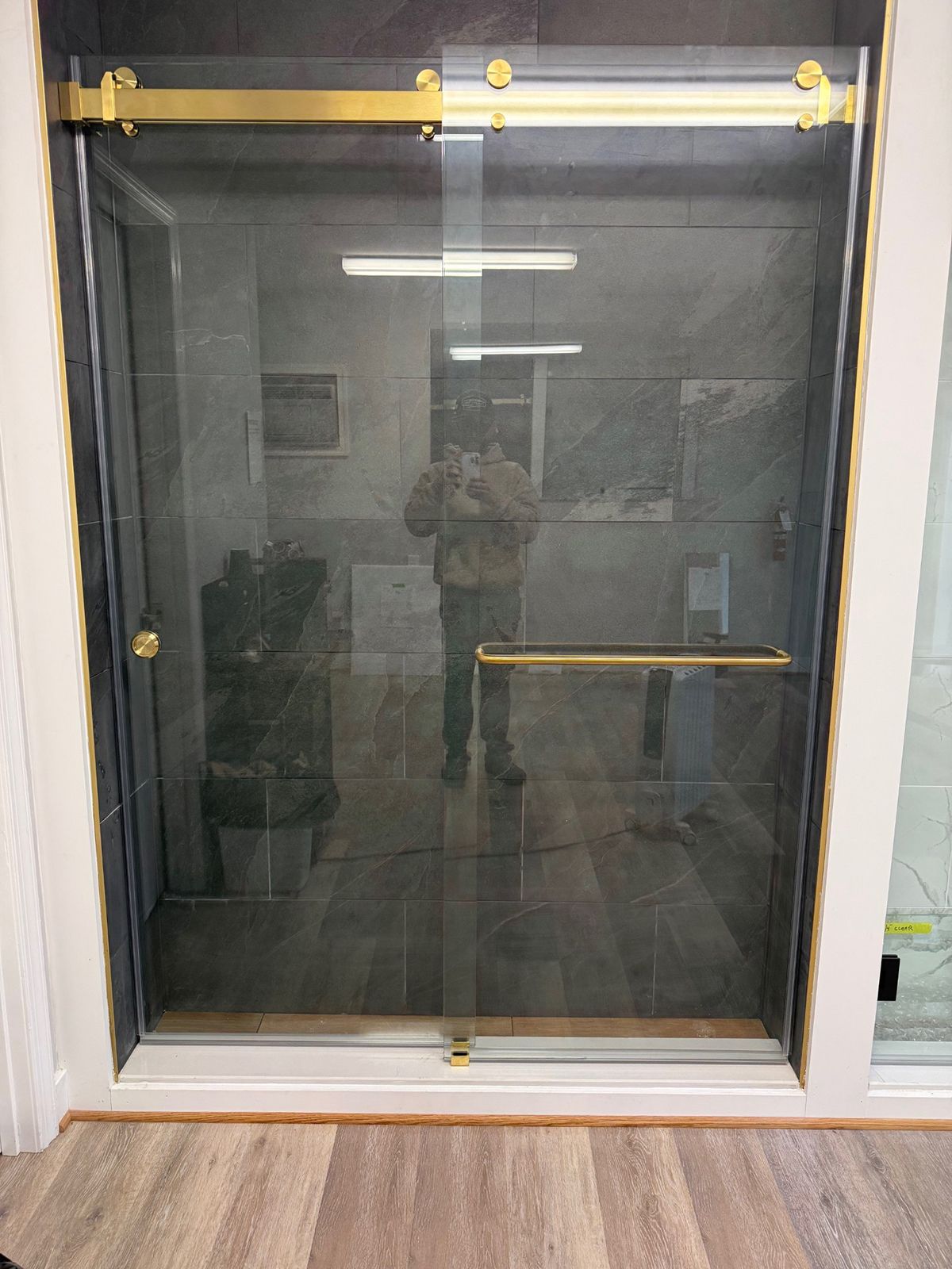 A glass shower door with gold hardware and a handle, installed in a home with wood-look flooring and dark tile walls.