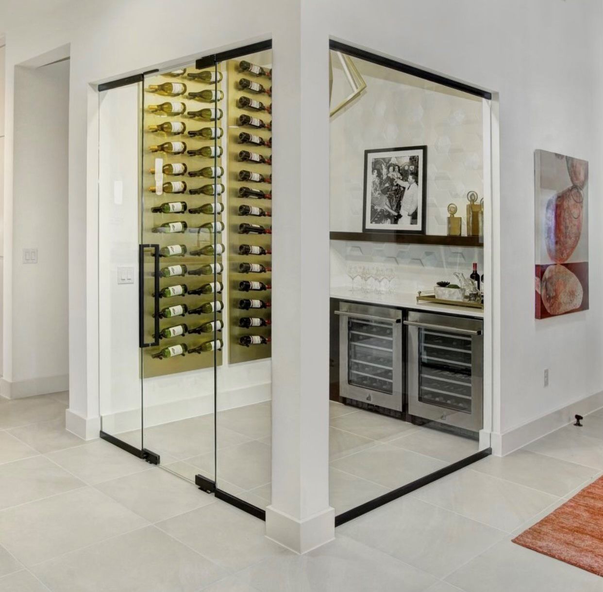 A modern glass-enclosed wine cellar with wall-mounted racks, dual refrigerators, and a bar counter in a minimalist room.