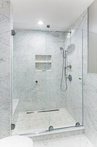 A walk-in shower with a glass sliding door, white marble walls, a recessed shelf, and chrome shower fixtures.