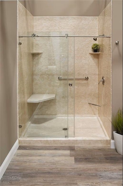 A walk-in shower with beige tile walls, a glass sliding door, built-in corner shelves, a corner seat, and safety grab bars.
