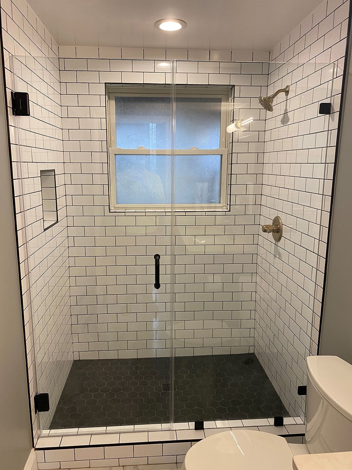 A walk-in shower with white subway tile walls, a dark stone floor, a window, and a glass door next to a toilet.