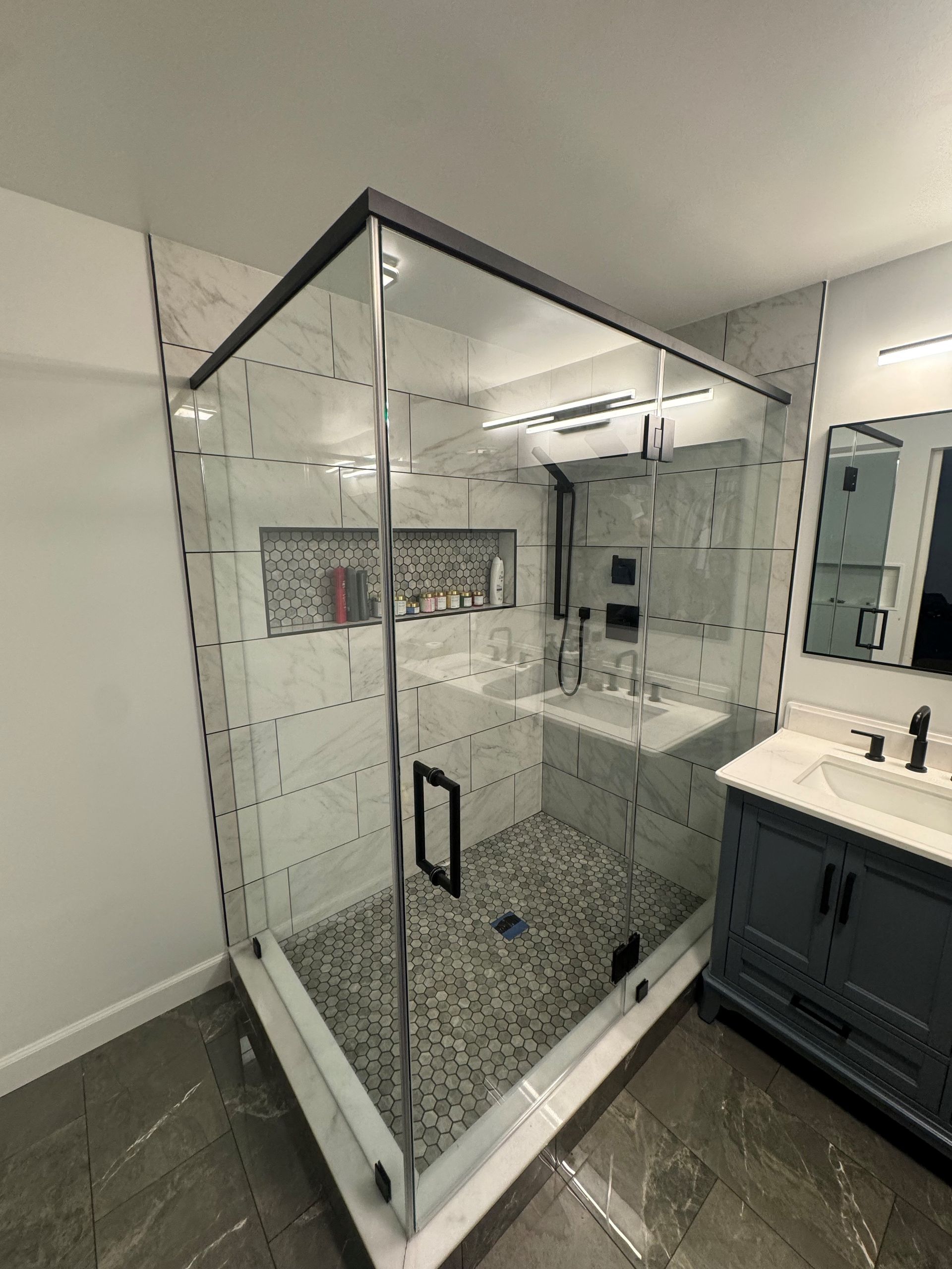 A corner glass shower enclosure with marble tiles, a pebble floor, and a built-in recessed niche in a modern bathroom.