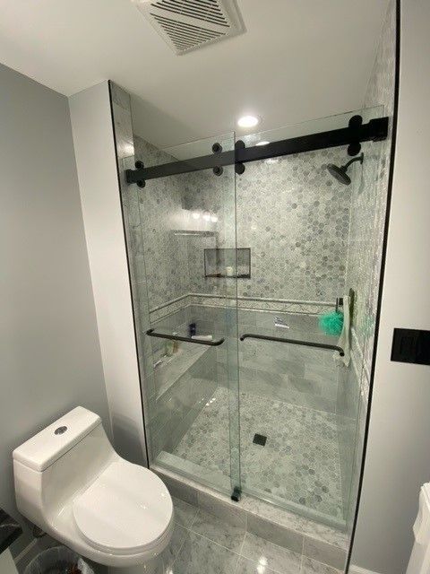 Bathroom with glass shower door, toilet, and window with blinds. Sunlight streams in.