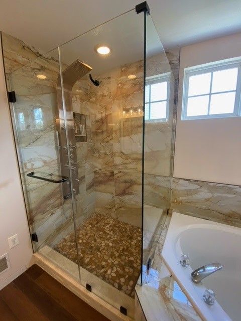 Modern bathroom with glass shower enclosure, tan marble walls and pebble floor. Corner tub visible.