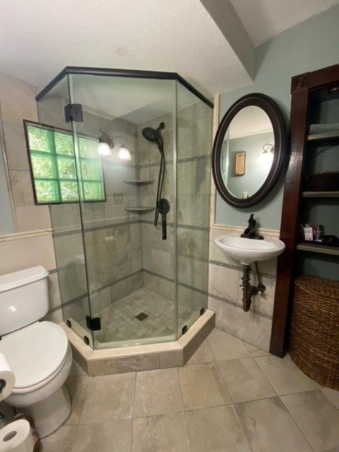Bathroom with a hexagonal glass shower, toilet, sink, mirror, and shelves. Beige tile floor and light green walls.