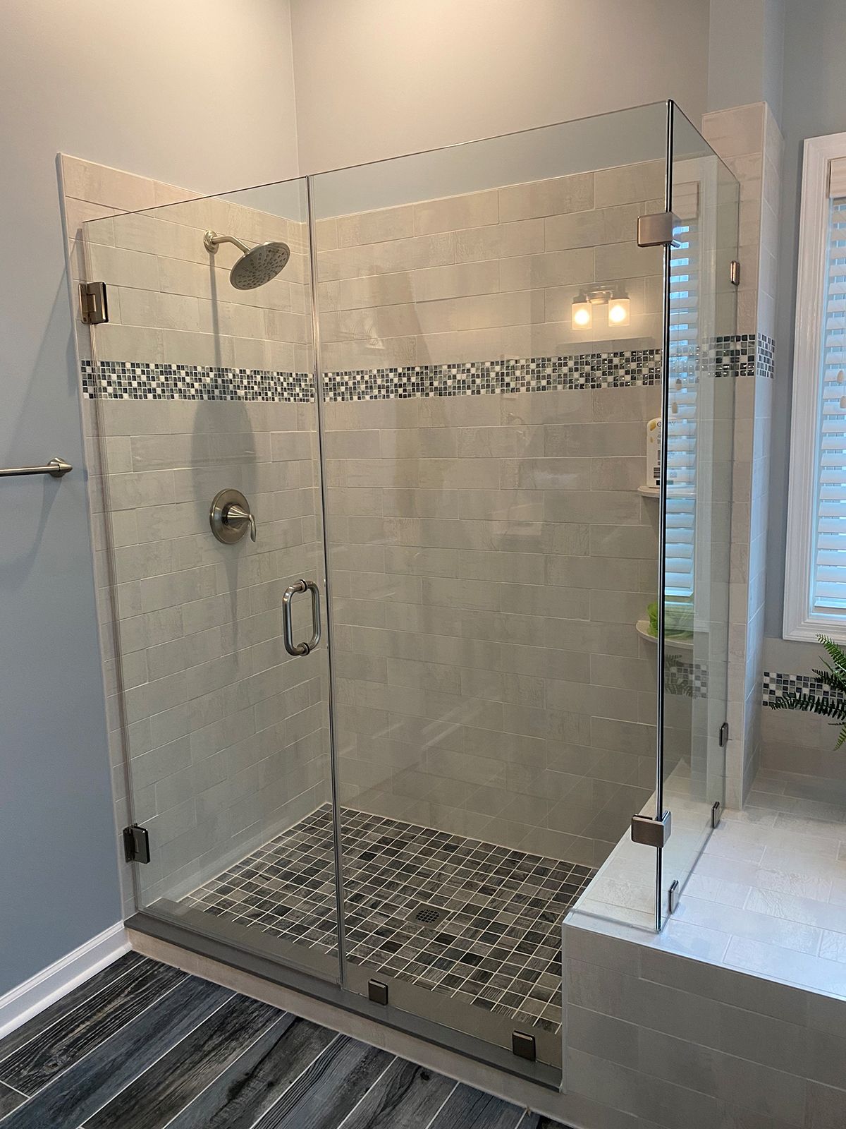 A walk-in glass shower with light tile walls, a decorative mosaic border, and a dark stone-tiled floor.