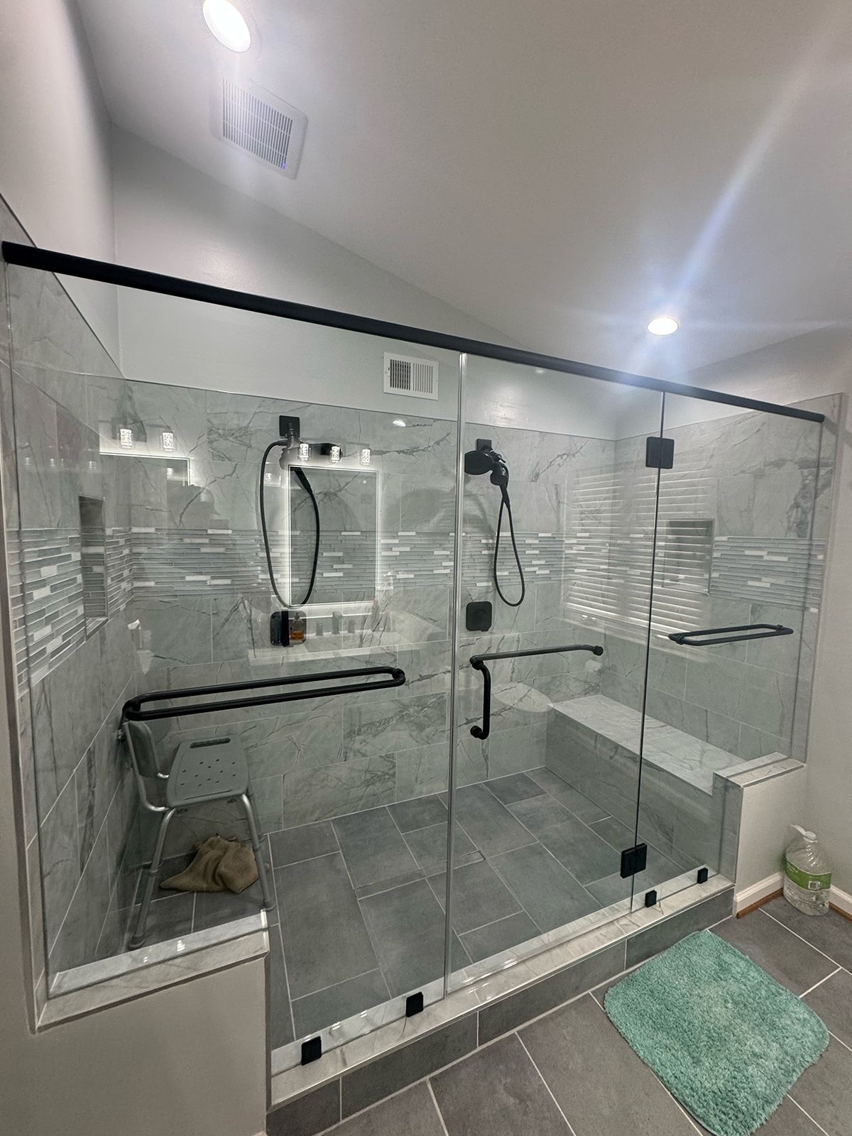 A large walk-in shower with a glass door, black hardware, gray tile walls and floor, a built-in bench, and shower seating.