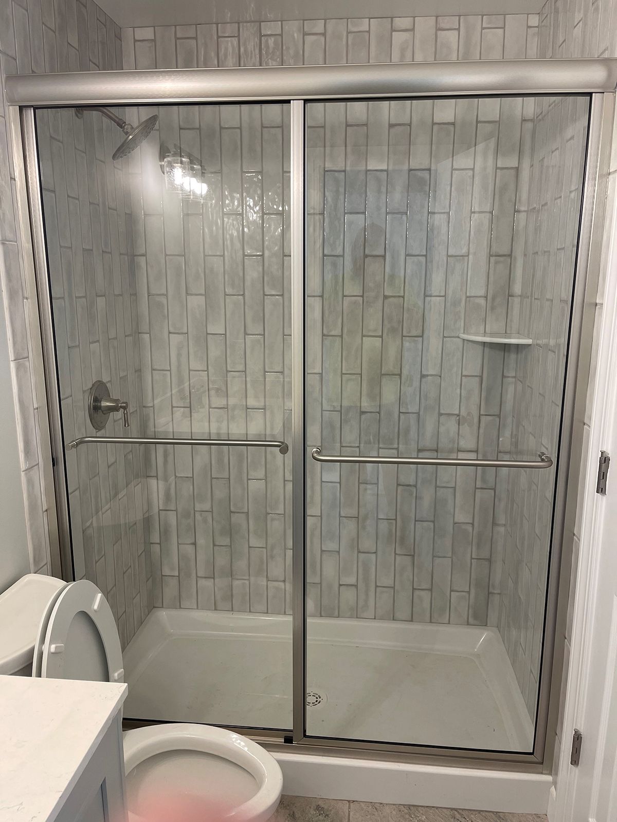 A shower stall with sliding glass doors and white vertical tile walls, situated next to a toilet in a bathroom.
