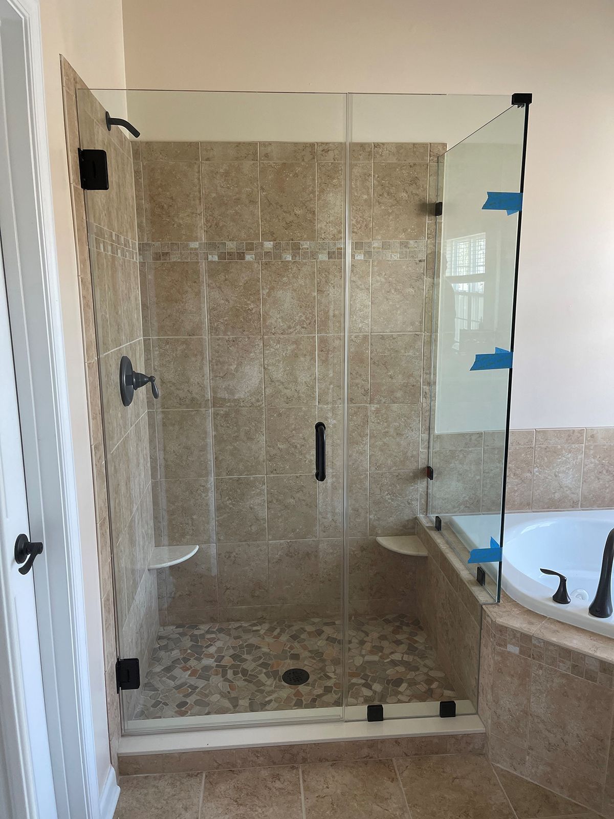 A glass shower enclosure with a dark faucet, handle, and hinges, set against tan tiled walls and a stone shower floor.