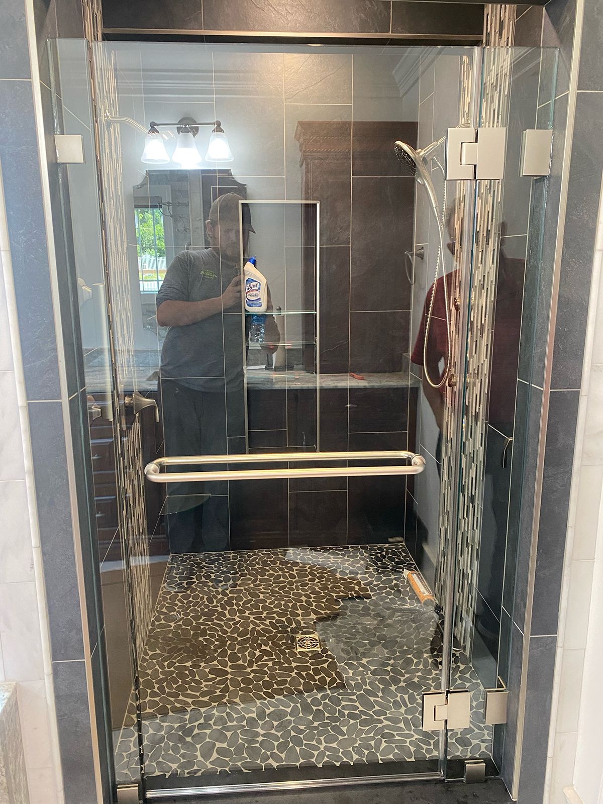 A glass shower door reflects a person holding a cleaning bottle in a bathroom with dark tile walls and mosaic floor tile.