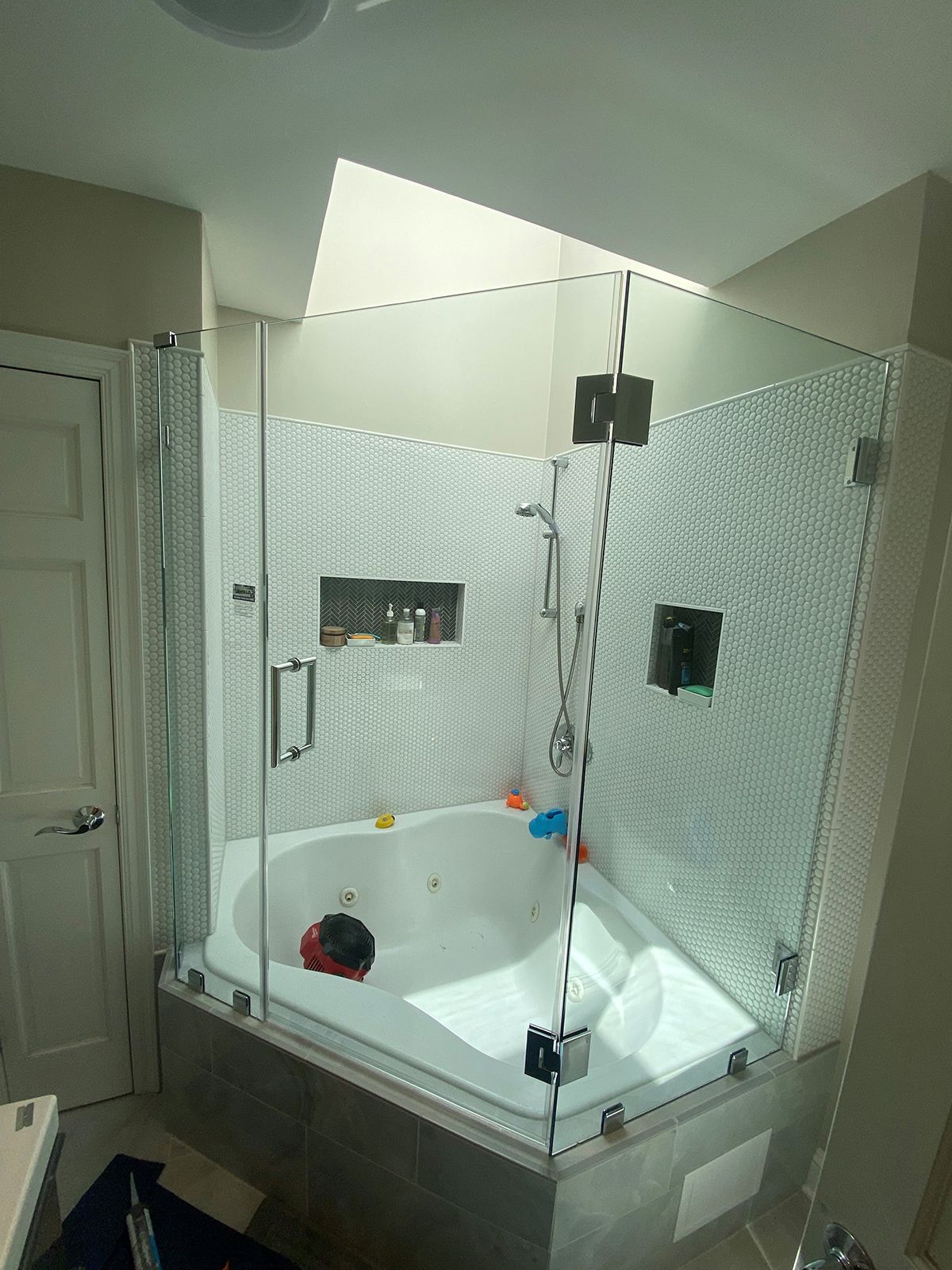 A corner soaking tub enclosed by a glass shower wall with mosaic tile surround and a recessed shelf.