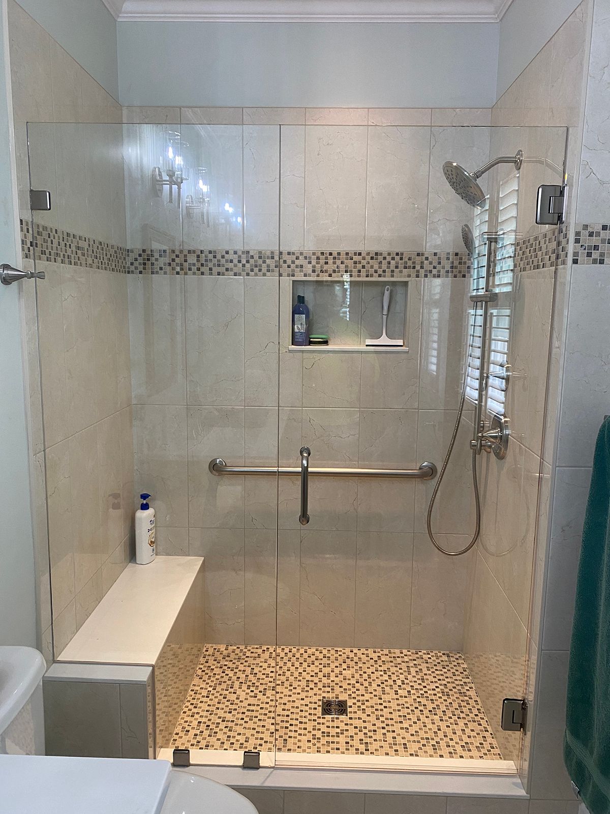 A walk-in shower with beige tiled walls, a built-in bench, pebble-style flooring, a grab bar, and a glass door.