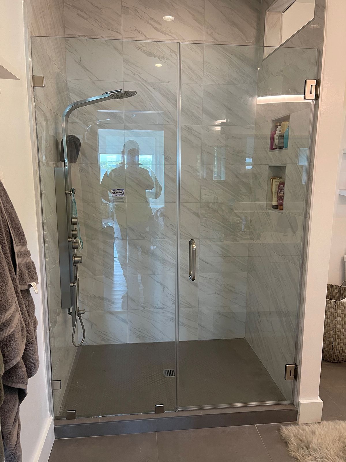 A glass-enclosed shower with marble-style tile walls, stainless steel fixtures, and built-in shelving.