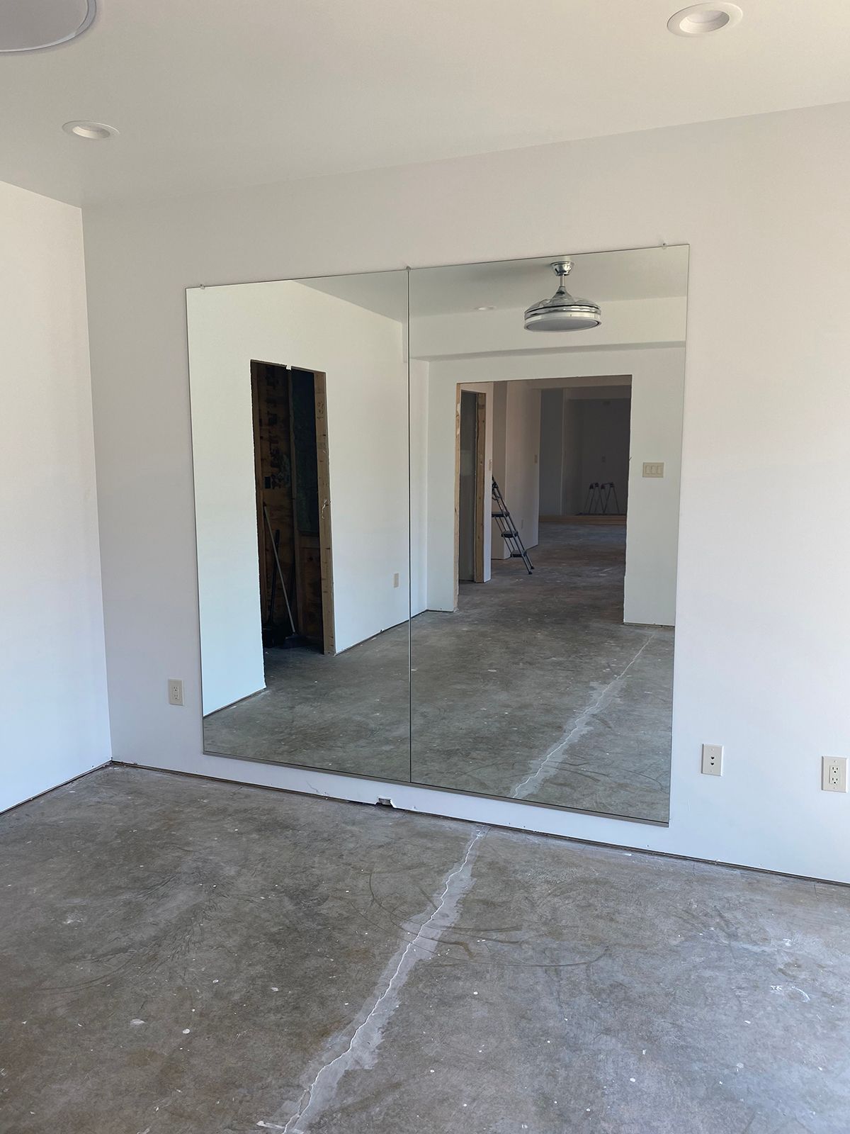 An unfinished room with concrete floors, white walls, and a large wall-mounted mirror reflecting an adjacent hallway.
