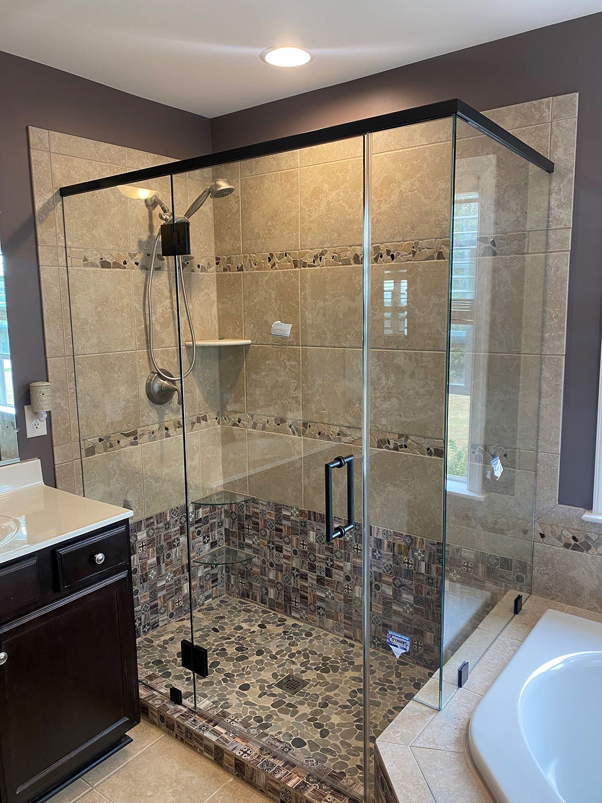 A corner walk-in shower with a black-framed glass enclosure, stone tile walls, and a pebble-stone floor.