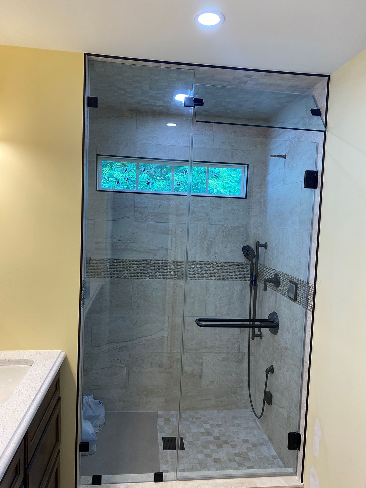 A walk-in shower with a glass door, black hardware, a horizontal grab bar, and stone-tiled walls in a bathroom.