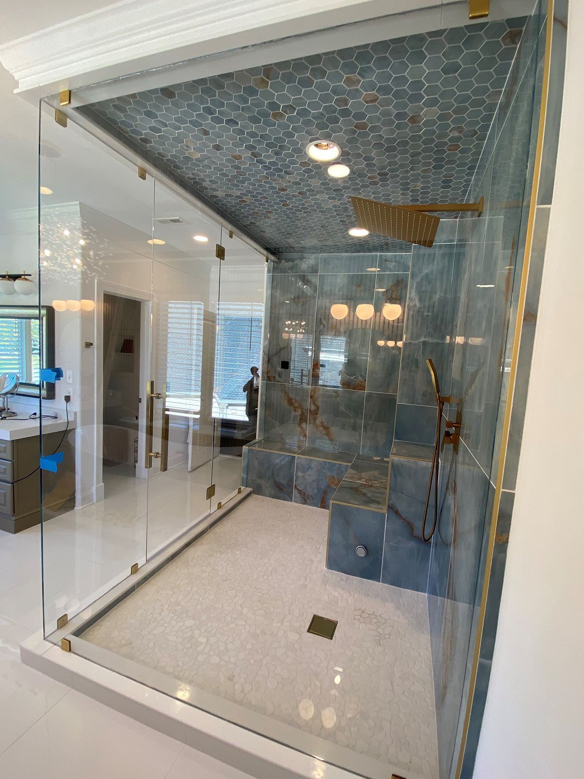 A modern walk-in shower with gold fixtures, blue-grey hexagonal ceiling tiles, stone walls, and a glass enclosure.
