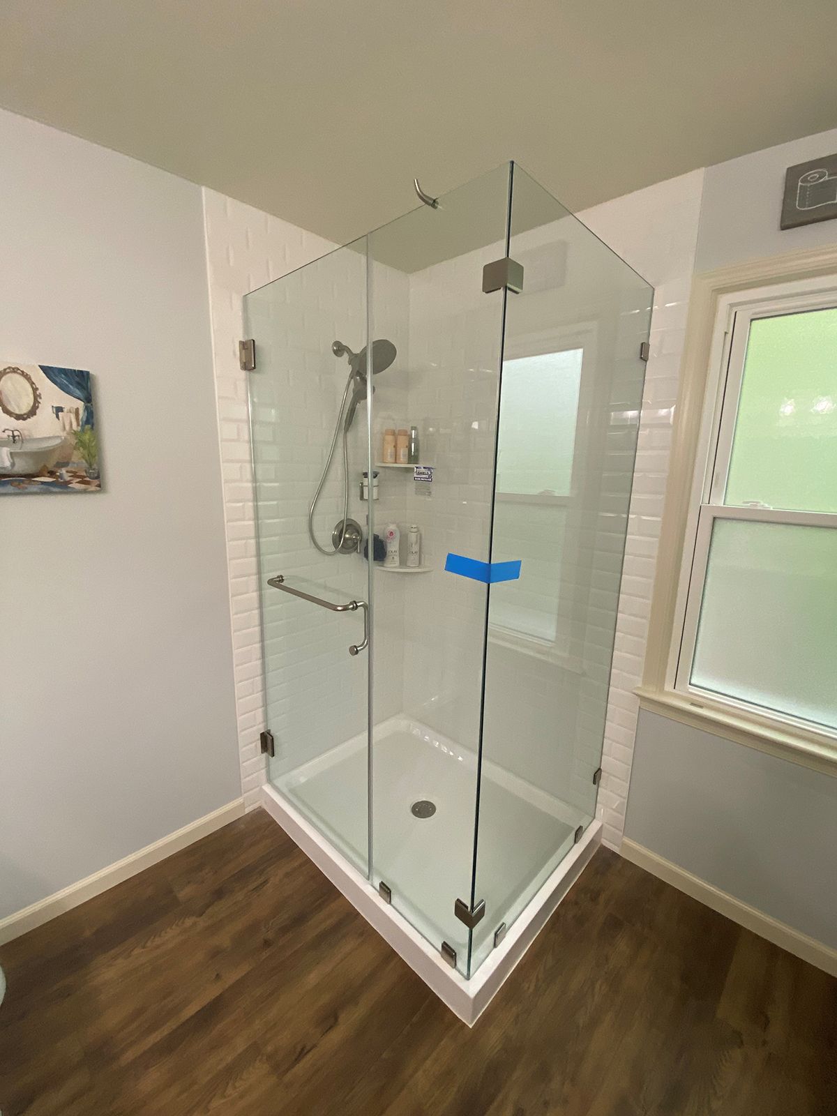 A glass-enclosed shower stall with a white subway tile surround, chrome hardware, and wood-look flooring.