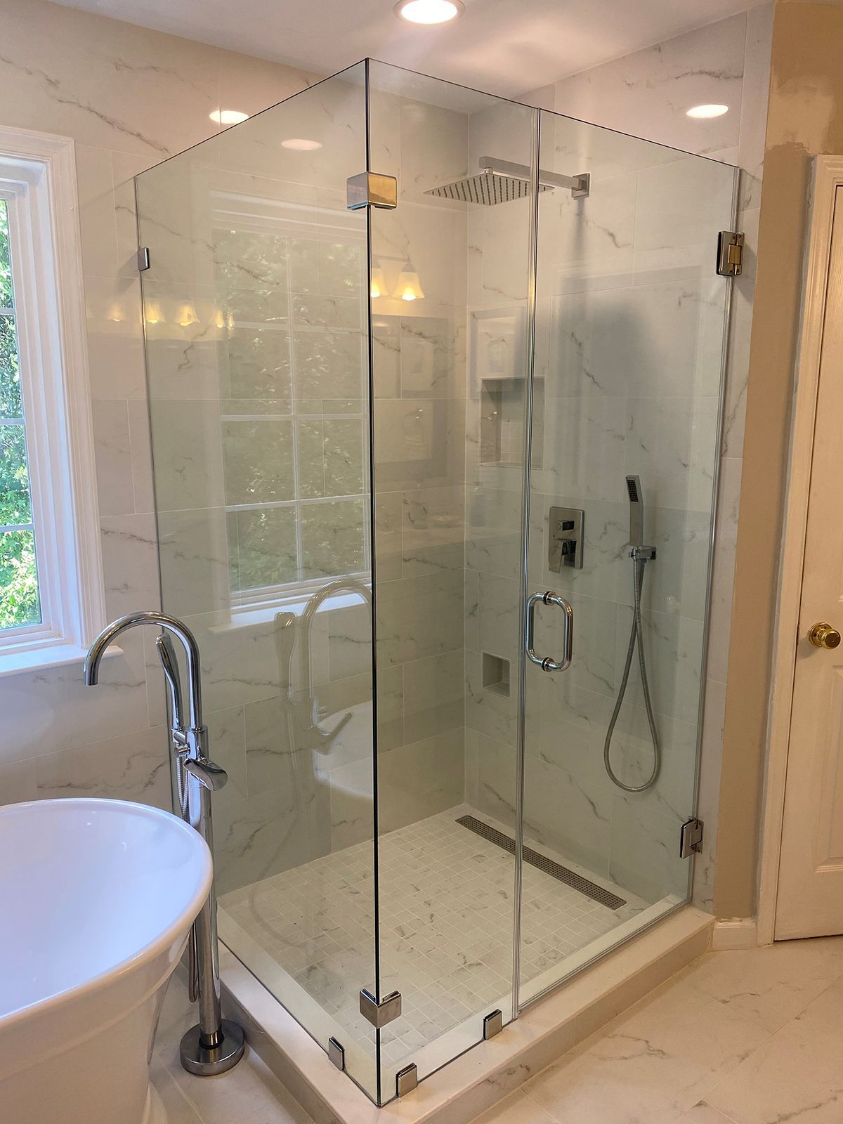 Modern glass shower stall with marble-tiled walls and floor, adjacent to a freestanding white bathtub in a bright bathroom.
