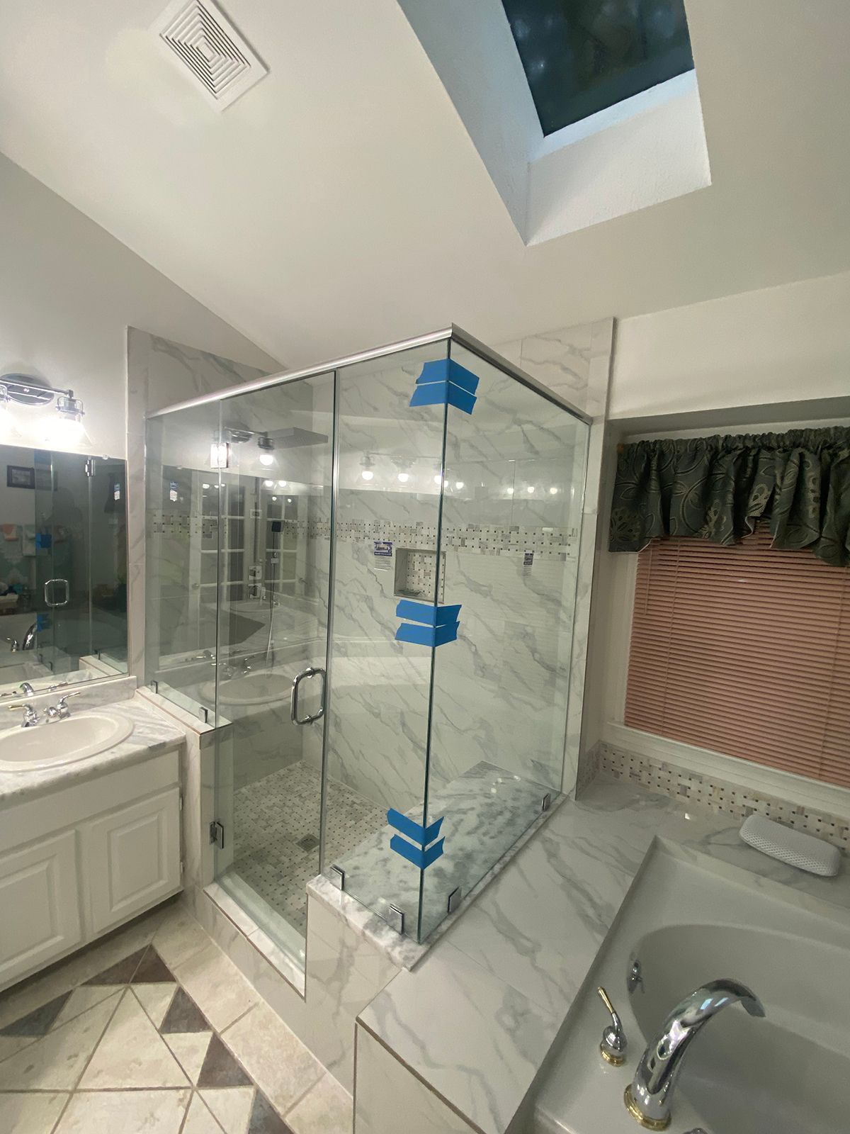 A newly installed glass shower enclosure with blue protective tape on the frame, set in a marble-tiled bathroom.