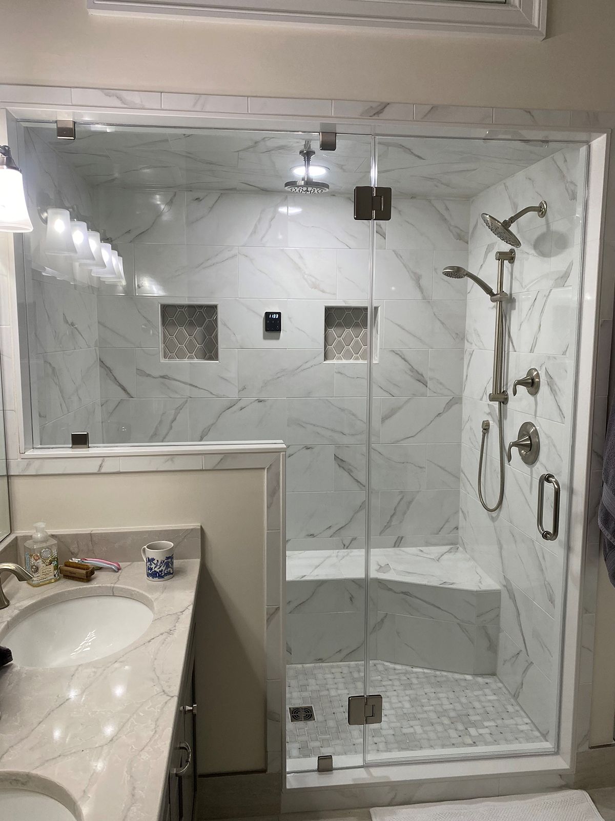 A walk-in shower with marble-look wall tiles, a built-in bench, glass door, and silver fixtures next to a bathroom vanity.