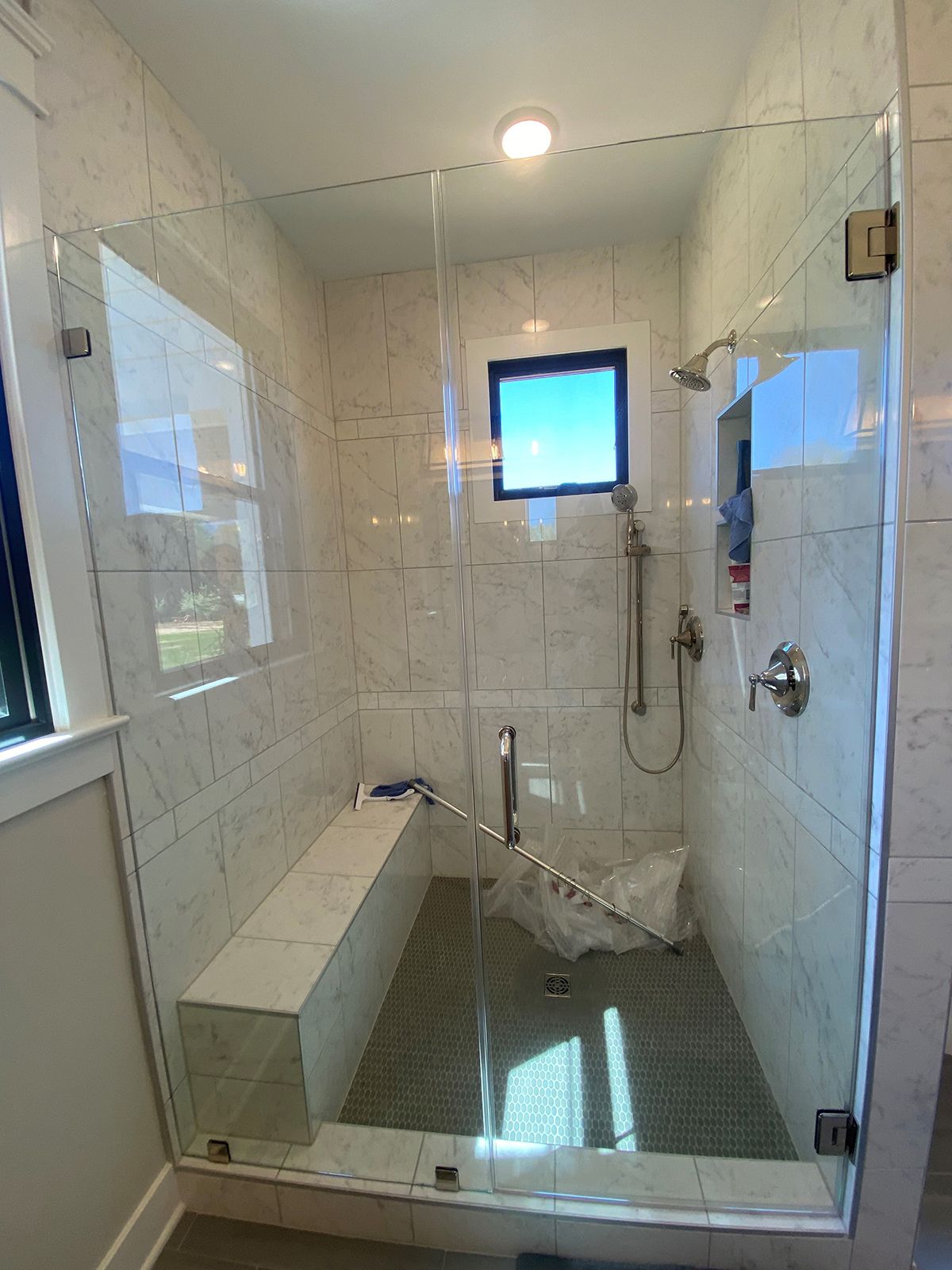 A walk-in shower with a glass door, marble-patterned walls, a built-in stone bench, and a small window above the showerhead.