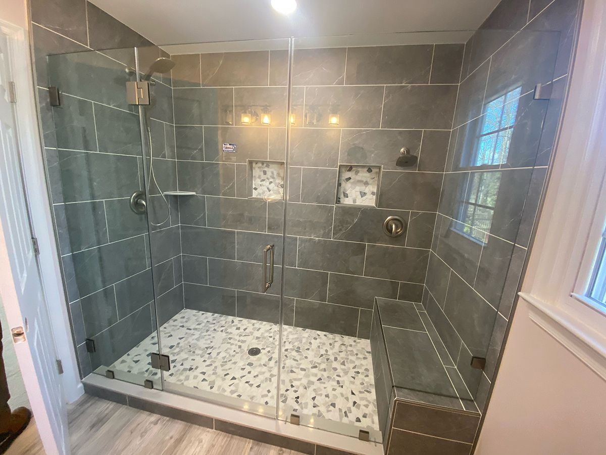 A modern glass-enclosed shower with gray tiled walls, pebble floor, built-in bench, two showerheads, and wall niches.