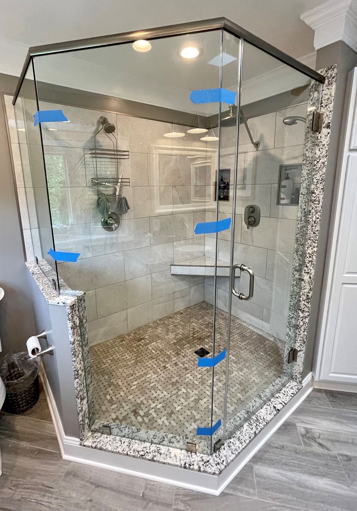 Corner shower enclosure with glass walls, speckled stone base and trim, and blue painter's tape on the glass seams.