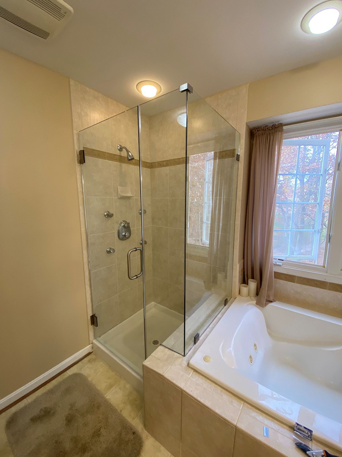 A bathroom featuring a glass shower stall next to a soaking tub, with light-colored tile and a nearby window with drapes.