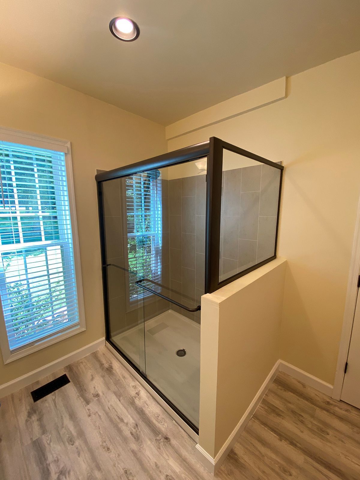 A corner shower with a glass enclosure and black frame, set in a room with light-colored walls and wood-patterned flooring.