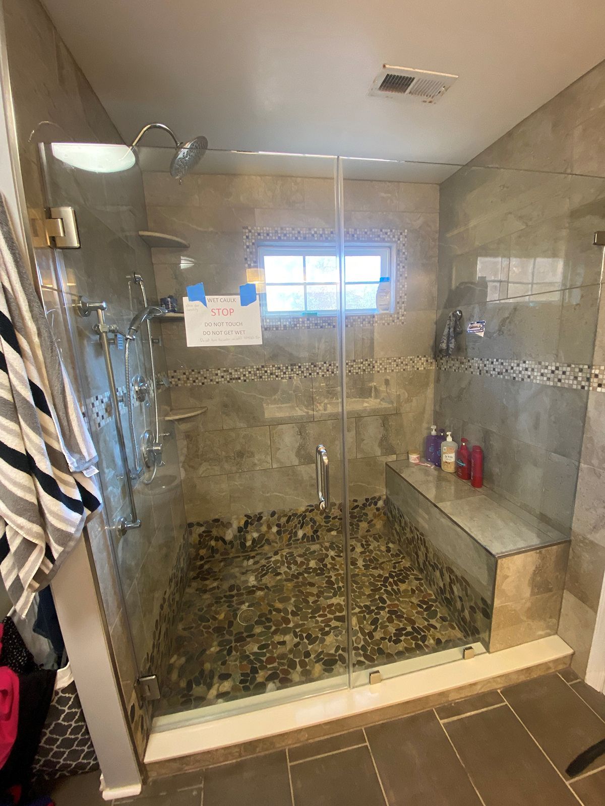 A walk-in shower with stone pebble flooring, a glass door, a built-in bench, and tiled walls under a recessed ceiling light.