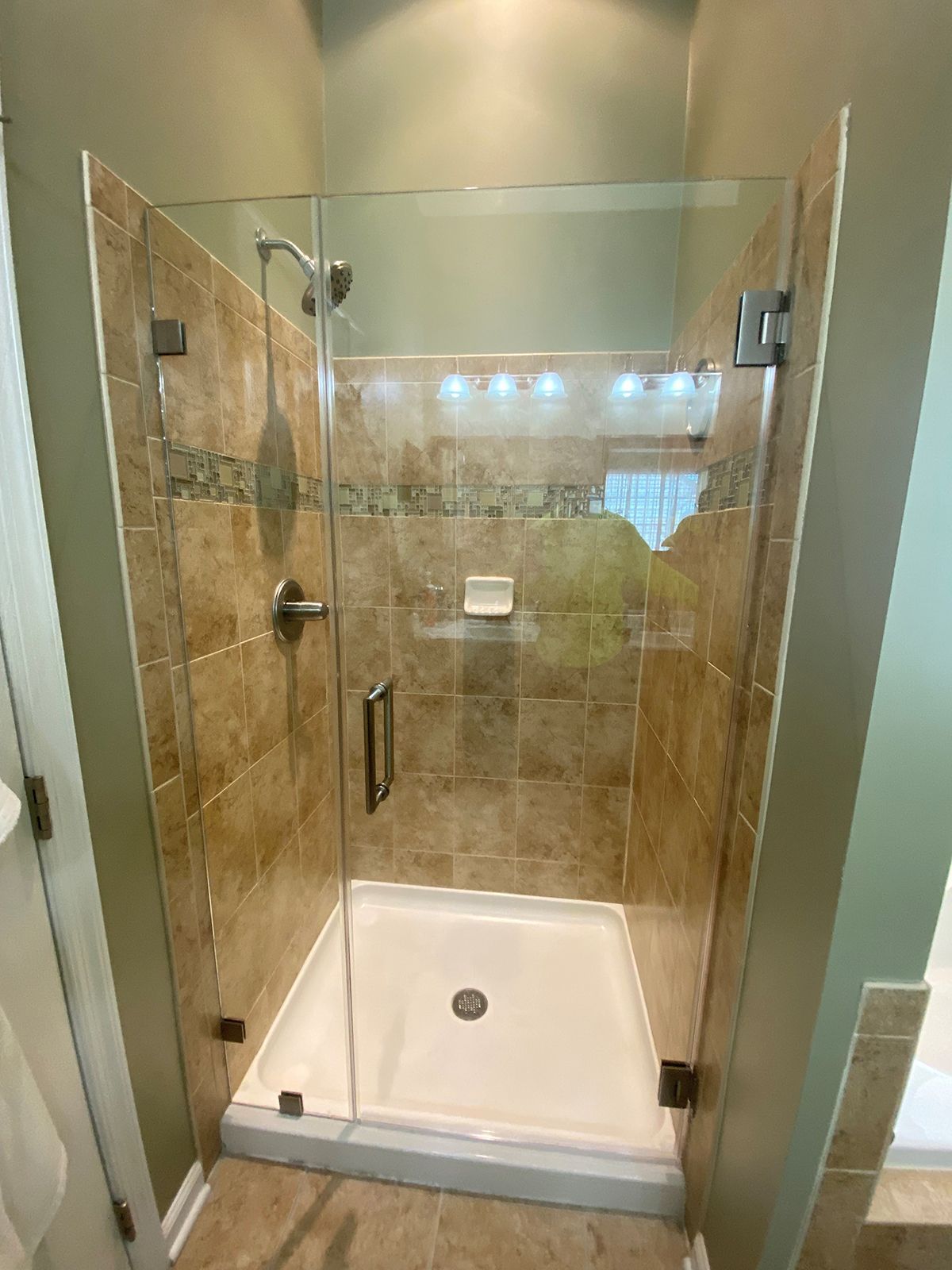 A walk-in shower with a glass door, tan tile walls, an accent border, a built-in shelf, and a white shower pan.