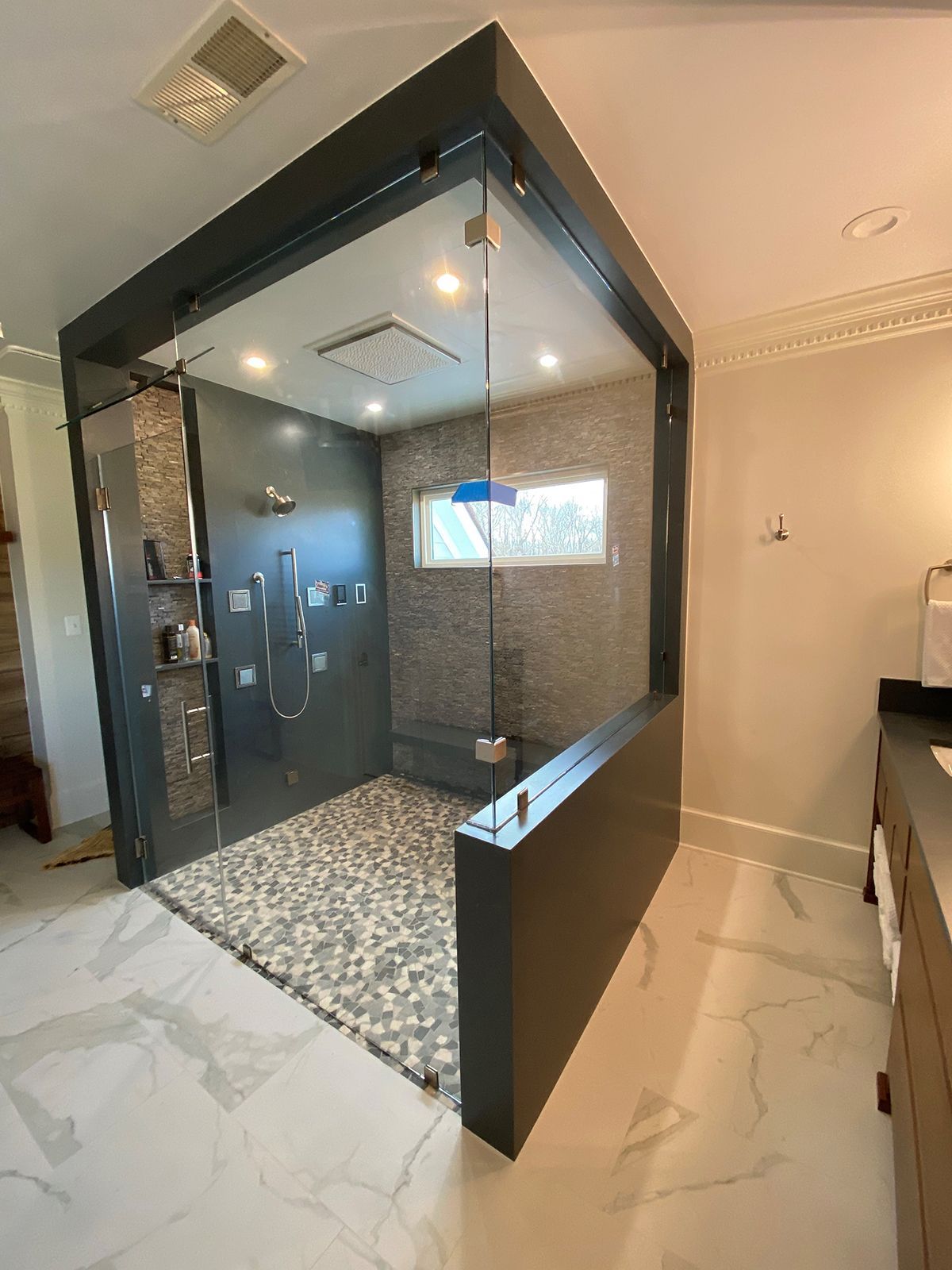 A modern glass-enclosed shower with pebble flooring and dark tile walls, set in a bright, marble-tiled bathroom.