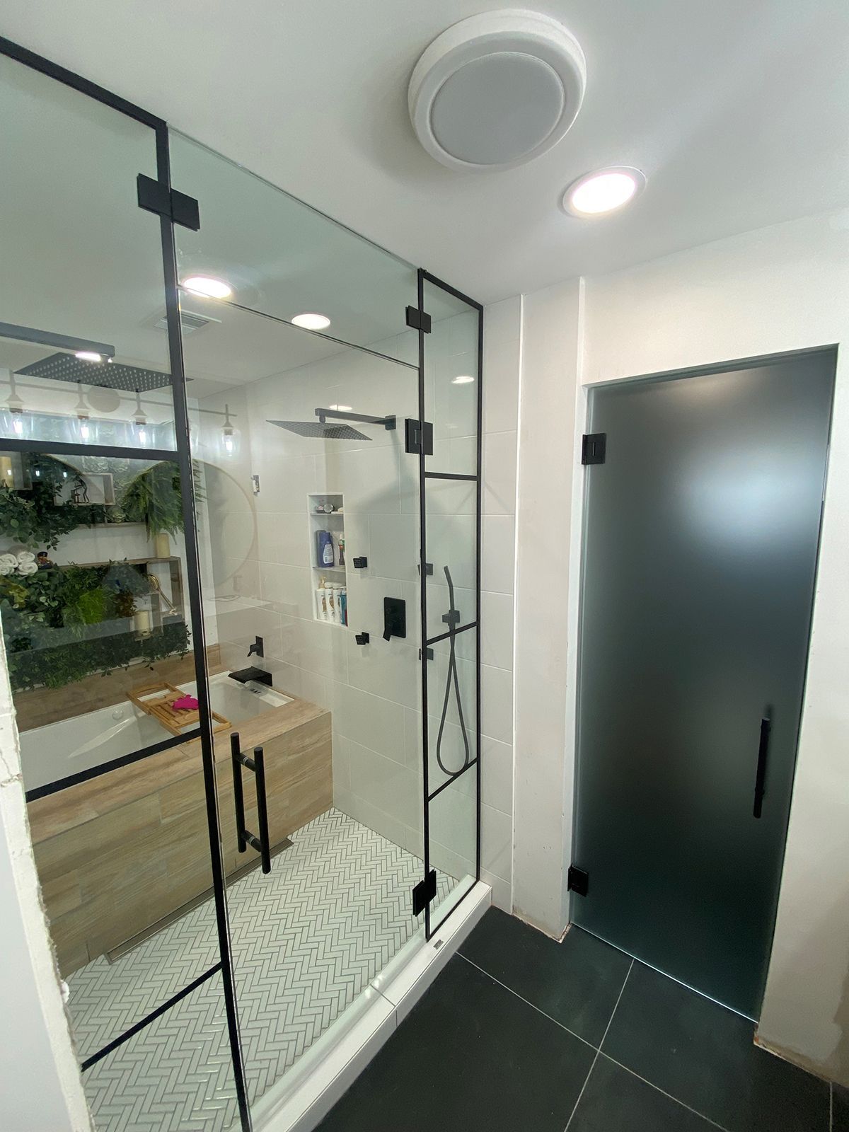 A walk-in shower with a black metal frame, glass walls, and a separate opaque black door in a modern bathroom.