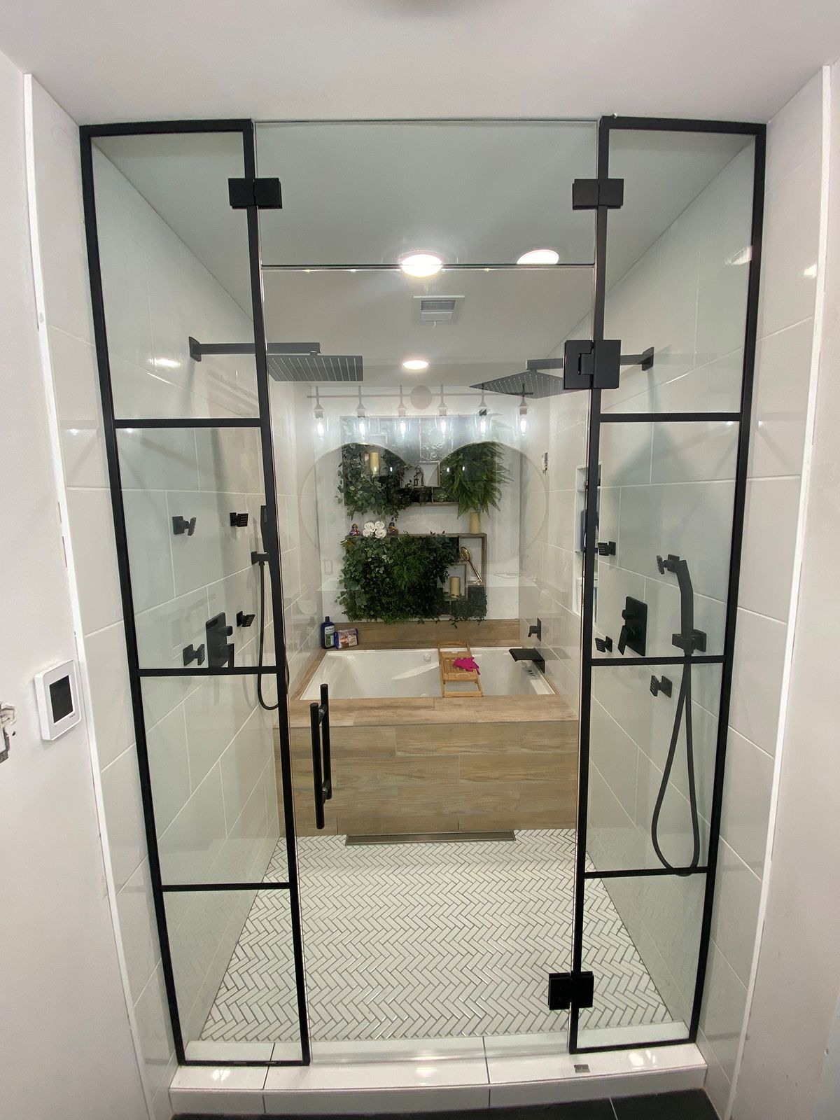 A modern shower with a black-framed glass door, white tiled walls, and a mosaic floor, looking toward a wooden vanity.