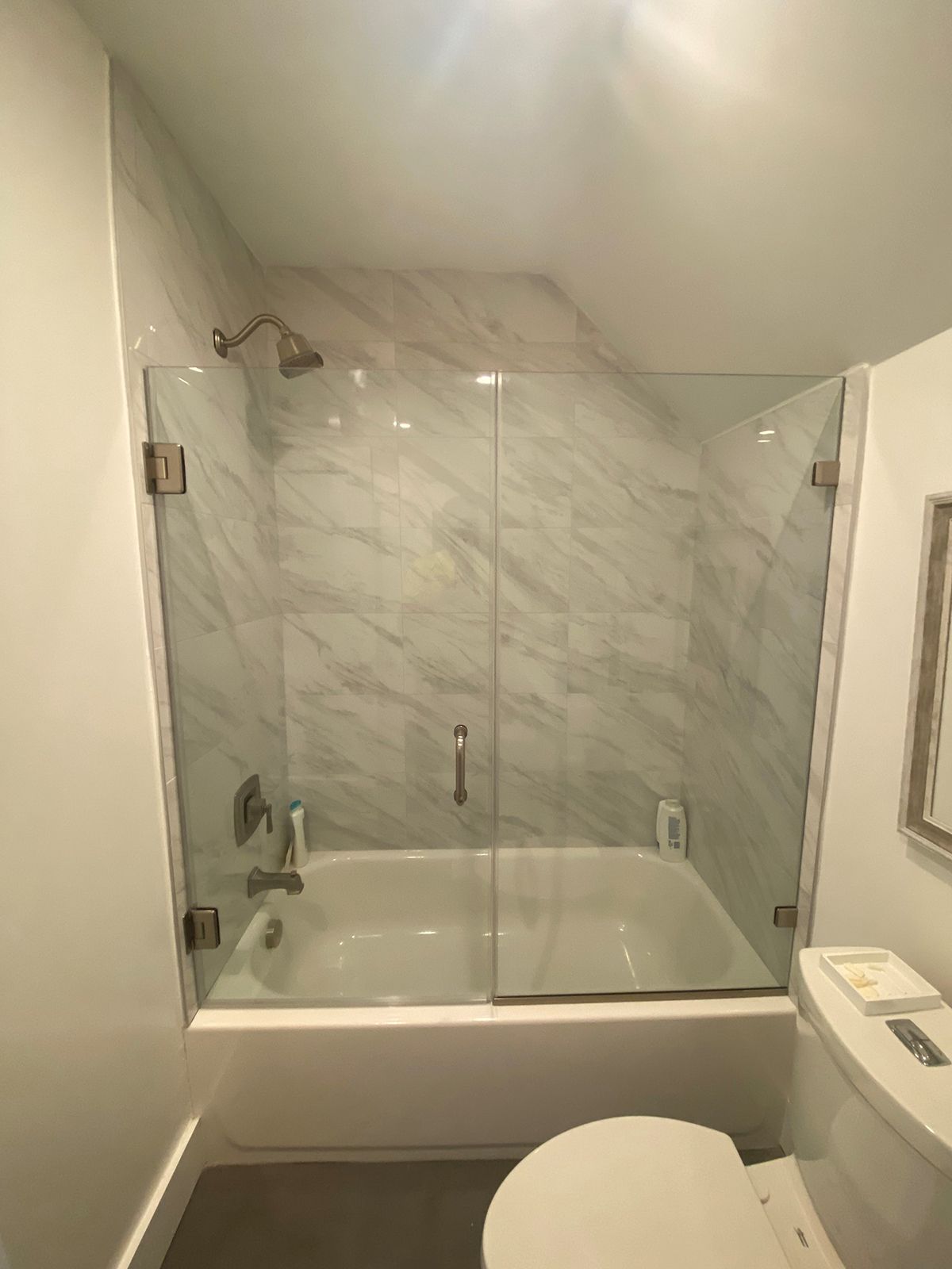 A bathroom featuring a white bathtub with a glass door, light marble-patterned tile walls, a showerhead, and a toilet.