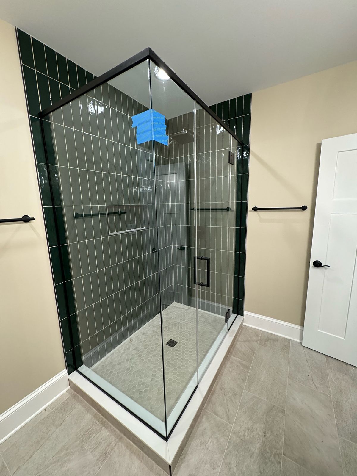 A modern glass-enclosed shower with dark green, rectangular wall tiles, a light pebble floor, and black trim and fixtures.