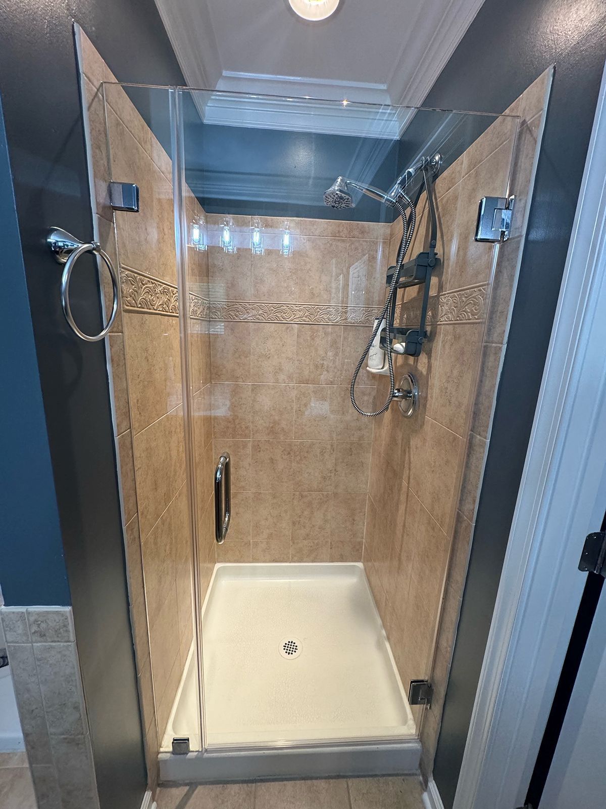 A walk-in shower with a glass door, tan tiled walls, a white floor pan, and a mounted silver shower head and controls.