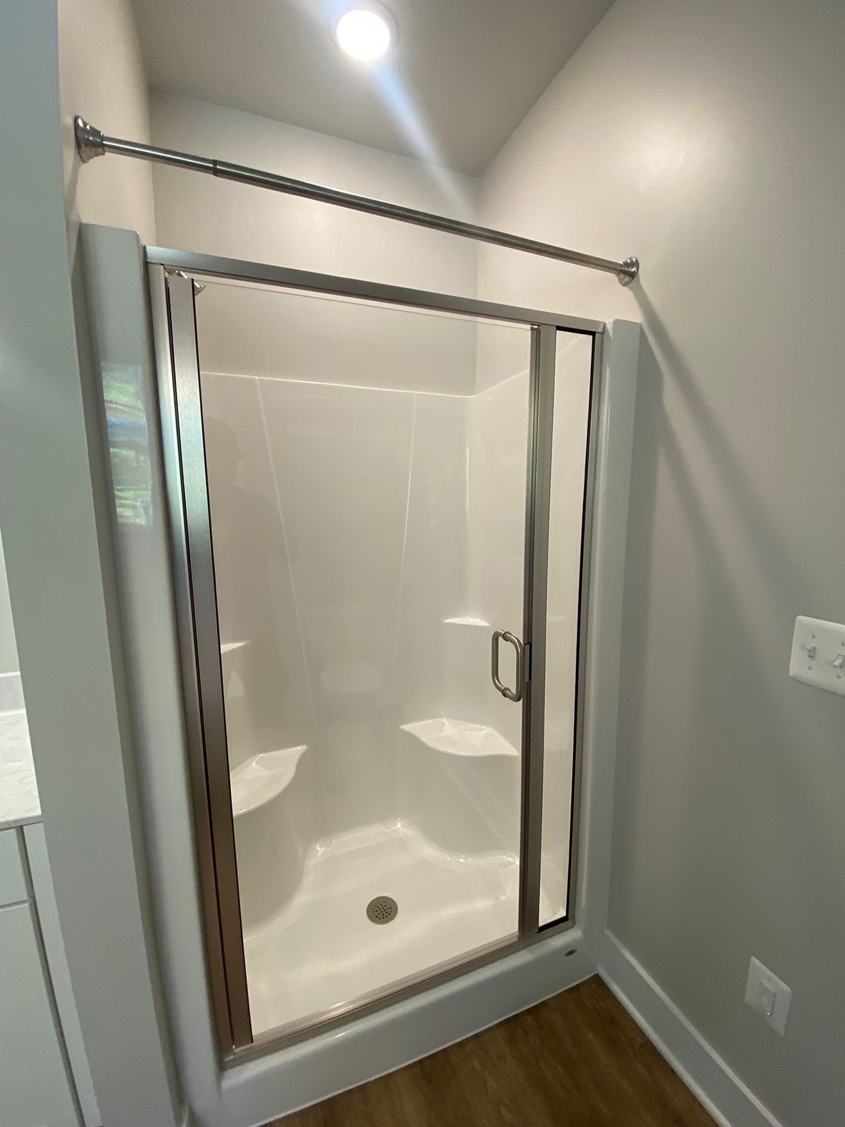 A corner shower stall with a white acrylic unit, glass door, and silver frame in a room with light grey walls.