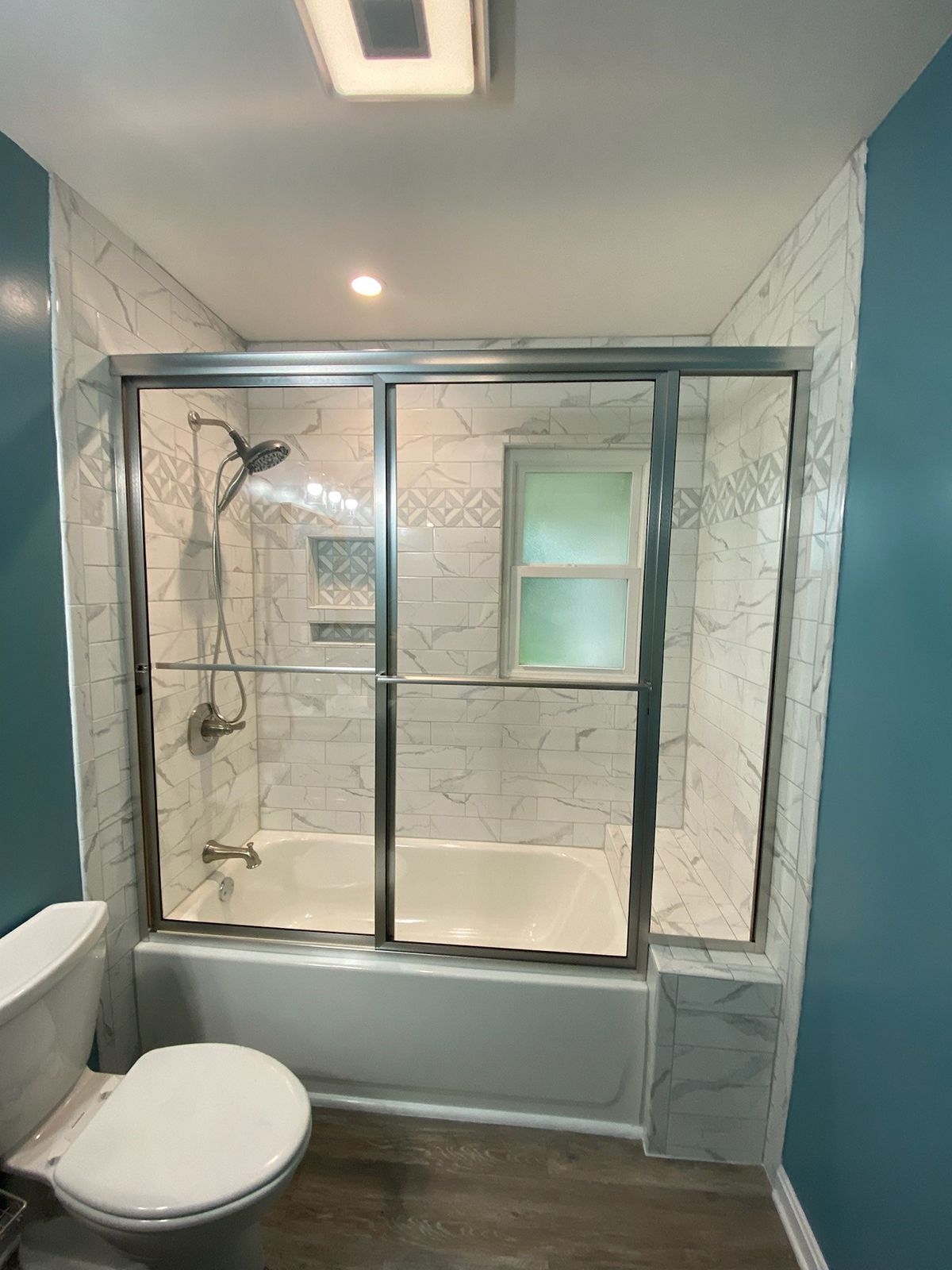 A bathroom featuring a white bathtub with sliding glass doors, marble-patterned tile walls, and blue walls.