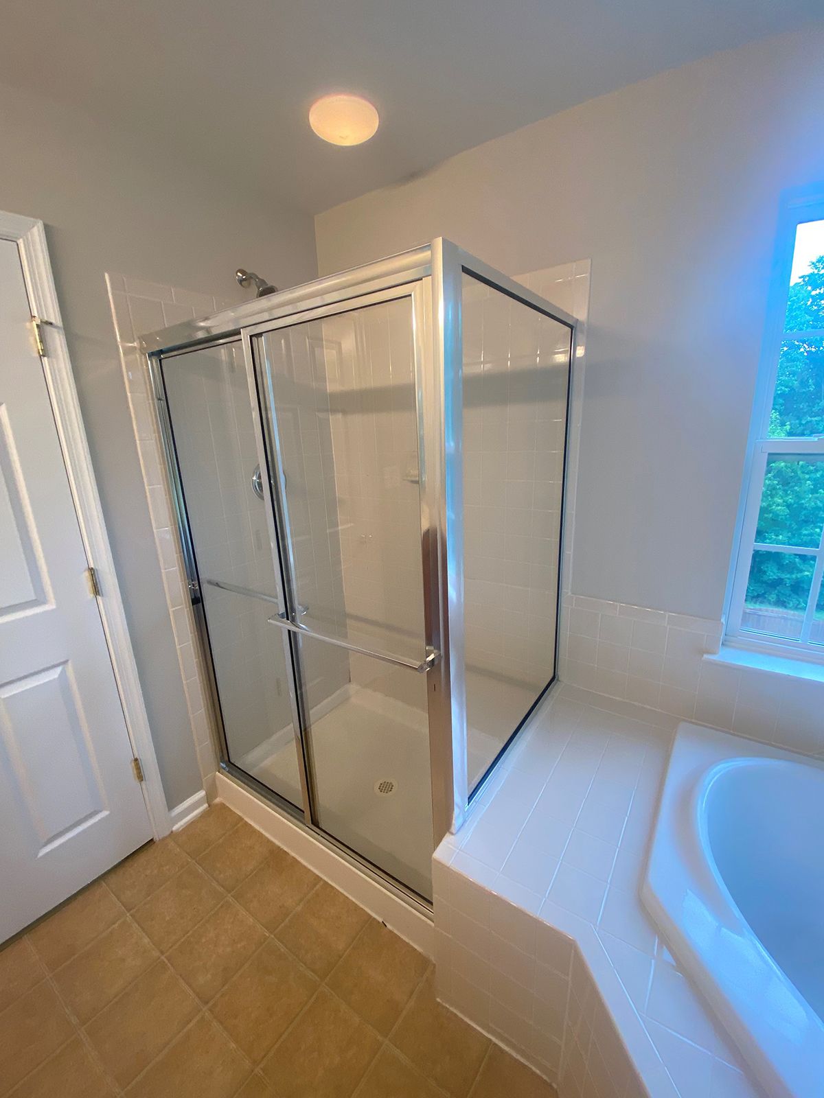 A bathroom corner featuring a glass-enclosed shower with a silver frame next to a white bathtub and beige tiled floor.