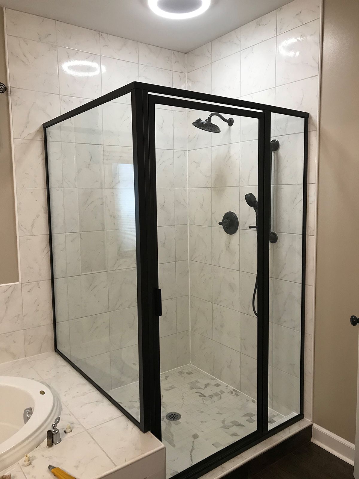 A glass shower enclosure with a black frame and white marble-patterned tile walls in a bathroom.
