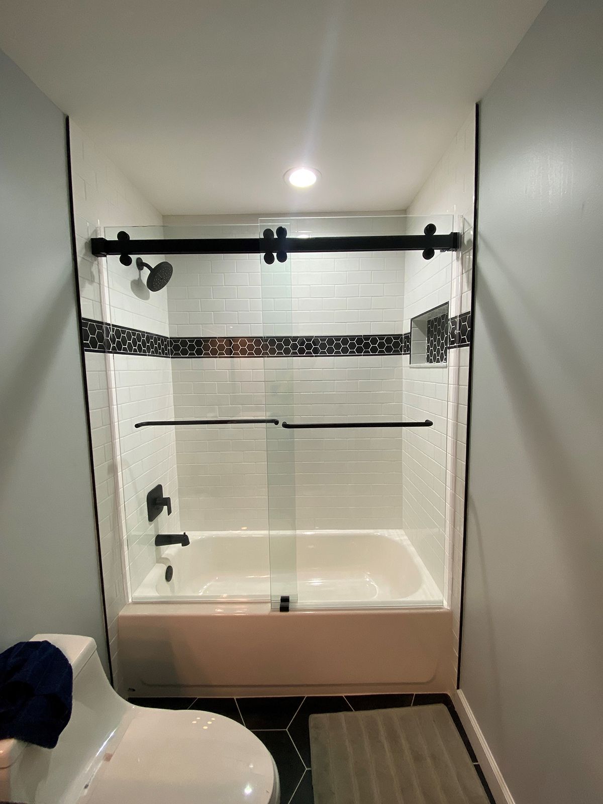 A white bathtub with a glass sliding shower door, black hardware, and white subway tile featuring a dark mosaic accent.