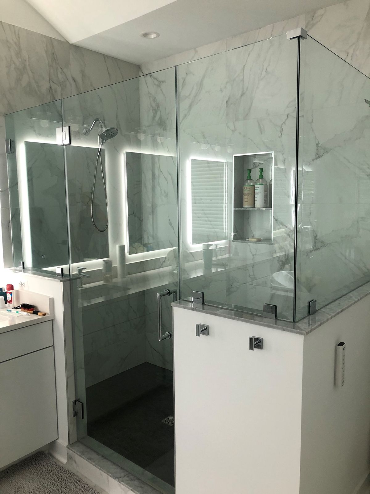 A walk-in shower with white marble walls, glass panels, a dark floor, and built-in shelving next to a modern vanity.