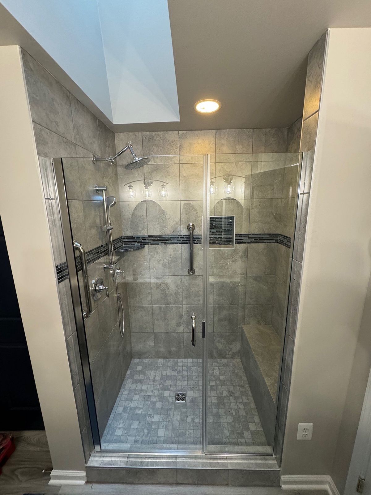 A modern walk-in shower with gray stone tile, a glass door, a built-in bench, and a handheld shower head fixture.